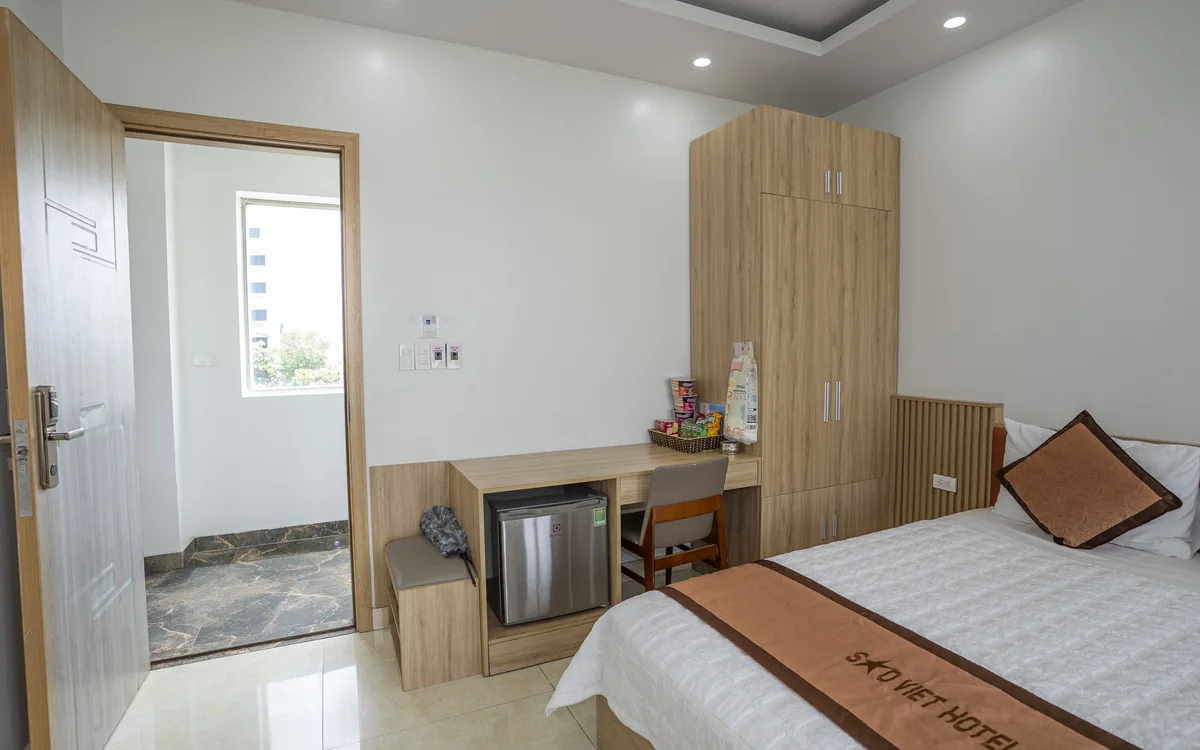 Giờ check in khách sạn – phòng giường đôi ga trắng nhìn từ cửa ra vào, tủ gỗ và minibar tại Sao Việt Hotel