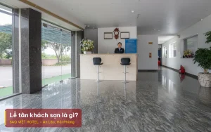 Lễ tân khách sạn là gì – ảnh minh hoạ nhân viên lễ tân đứng sau quầy đá ở sảnh khách sạn, kèm tiêu đề bài viết