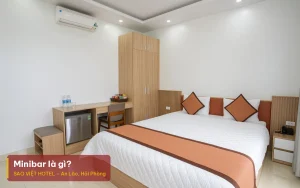 Minibar là gì – ảnh minh hoạ phòng khách sạn có tủ lạnh nhỏ dưới bàn, chai nước và giỏ đồ ăn nhẹ, kèm tiêu đề bài viết