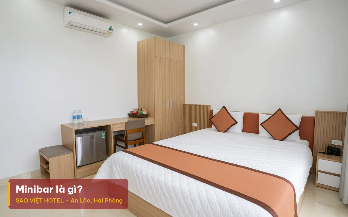 Minibar là gì – ảnh minh hoạ phòng khách sạn có tủ lạnh nhỏ dưới bàn, chai nước và giỏ đồ ăn nhẹ, kèm tiêu đề bài viết