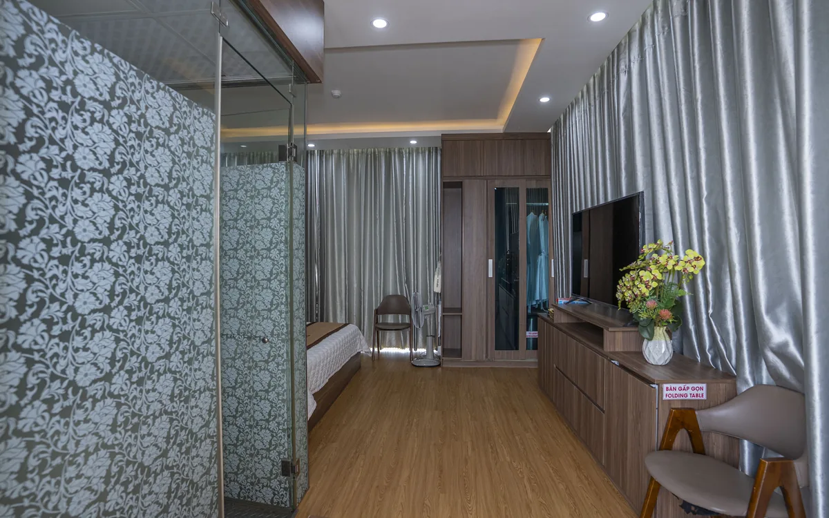 Phòng ốp gỗ có vách kính hoa văn, ghế gỗ, TV lớn và bình hoa vàng cạnh cửa sổ tại Sao Việt Hotel