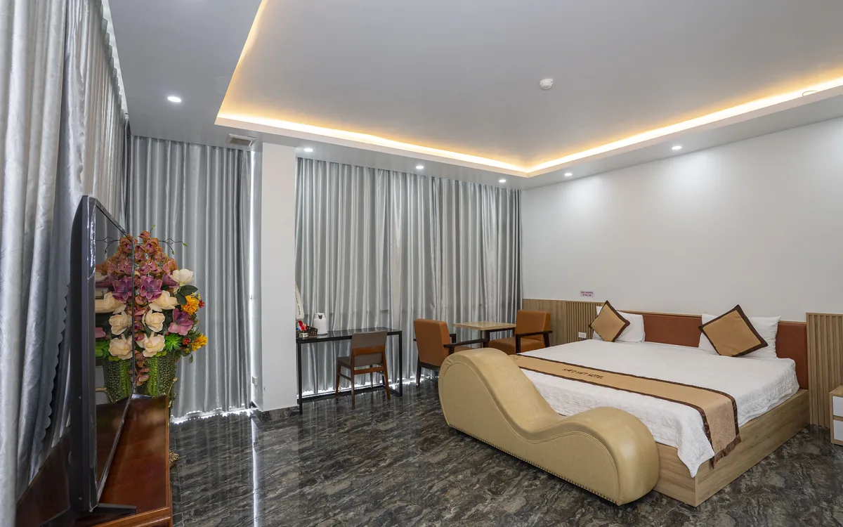 Phòng lớn đèn hắt trần với bình hoa ở tiền cảnh, giường đôi và ghế thư giãn màu be tại Sao Việt Hotel An Lão