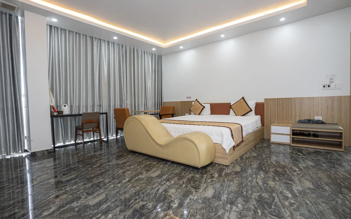 Phòng rộng sàn đá vân tối, giường đôi, ghế thư giãn màu be dáng cong và rèm xám toàn tường tại Sao Việt Hotel An Lão
