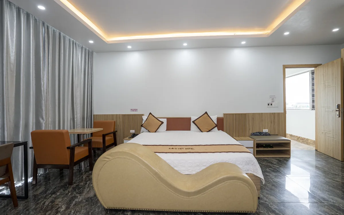 Phòng lớn sàn đá tối, đèn hắt trần, giường đôi, ghế thư giãn màu be dáng cong và cặp ghế bành màu cam tại Sao Việt Hotel
