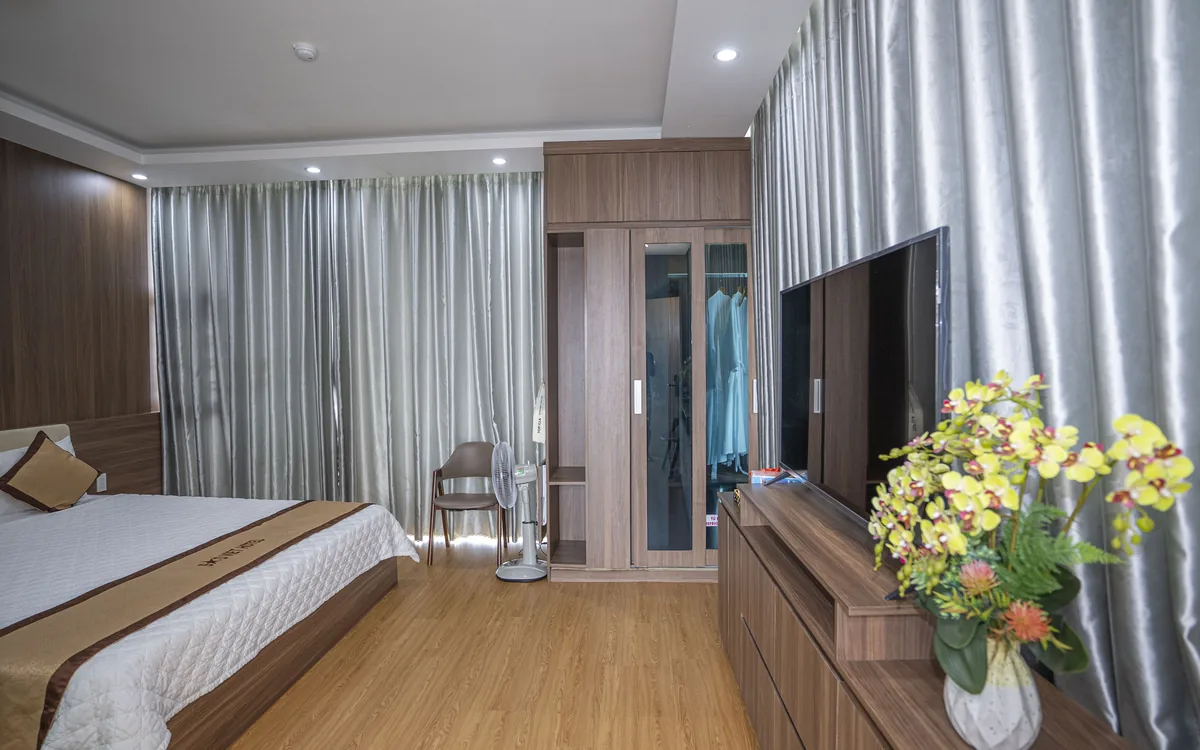 Phòng ốp gỗ sàn gỗ với TV lớn, kệ gỗ, bình hoa vàng và rèm xám tại Sao Việt Hotel An Lão
