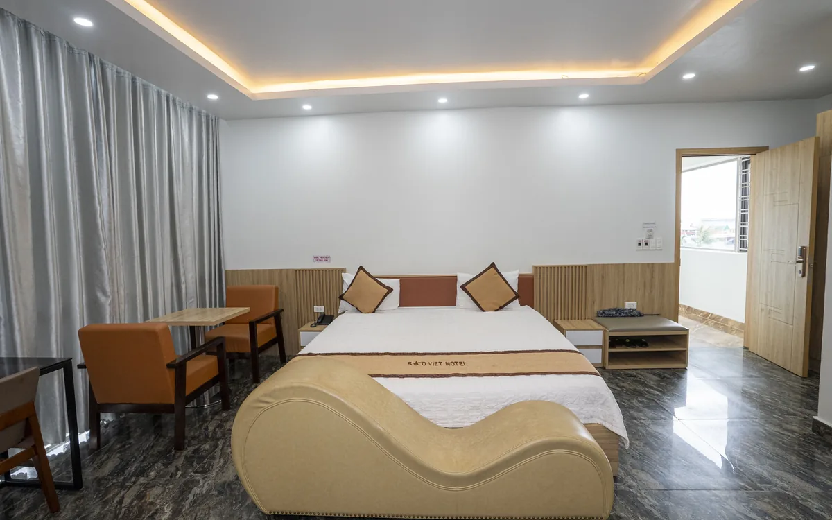Phòng lớn sàn đá vân tối với giường đôi, ghế thư giãn màu be, bàn làm việc và cặp ghế bành cạnh rèm xám tại Sao Việt Hotel