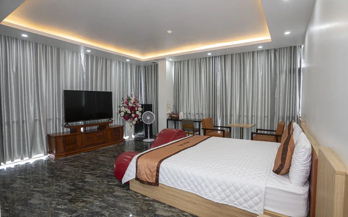 Phòng diện tích lớn sàn đá vân tối, TV trên kệ gỗ, giường đôi ga trắng và rèm xám toàn tường tại Sao Việt Hotel